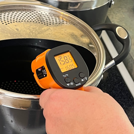 Infrared hand-held thermometer gun being used to check the temperature of a liquid in a pot on a stove.