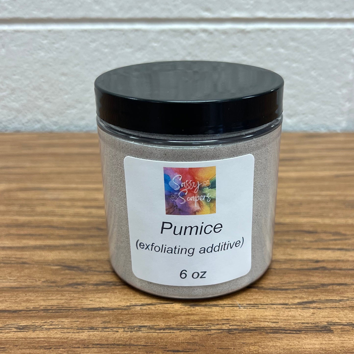 Round, 6 oz clear plastic container of pumice on wood grain table.