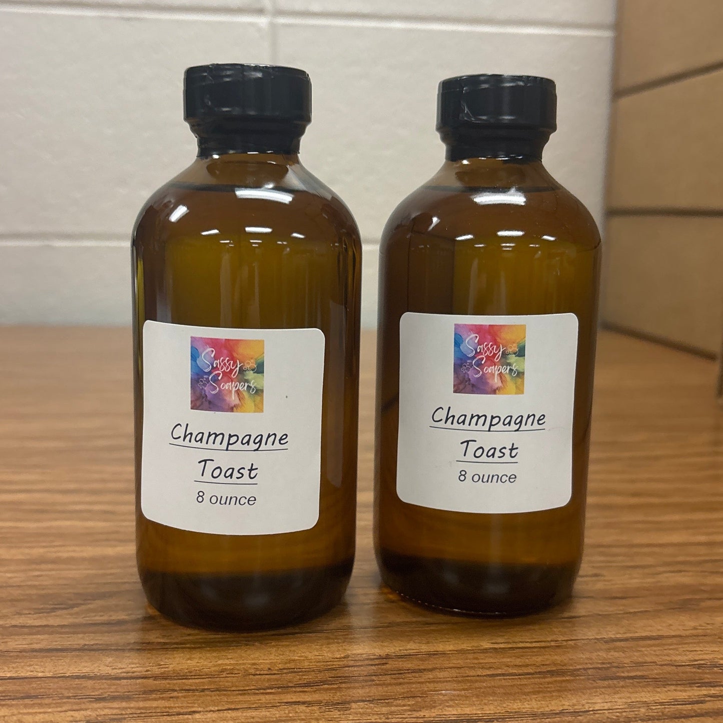 Two bottles of 'Champagne Toast' fragrence oils on a wooden surface with a tiled wall background.