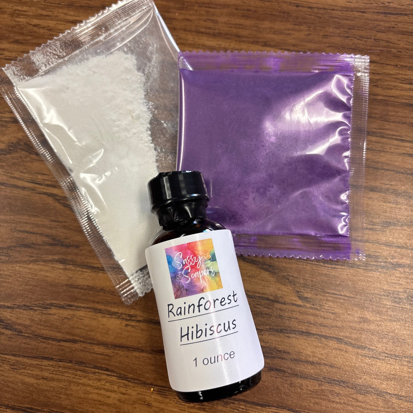 Bottle of Rainforest Hibiscus tea fragrence oil, purple bag, and white powder on a wooden surface