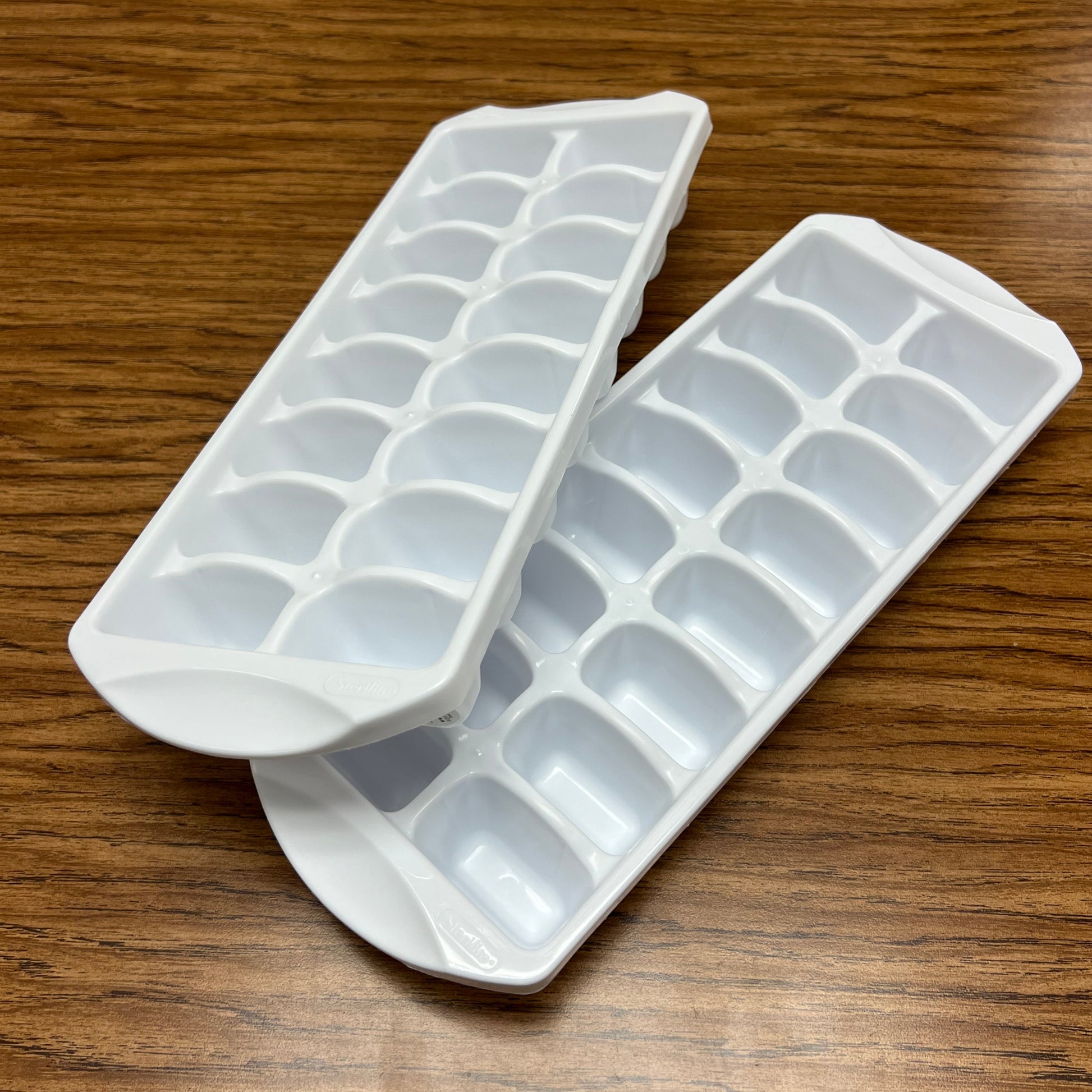 Pair of white Sterilite brand ice cube trays on woodgrain background.