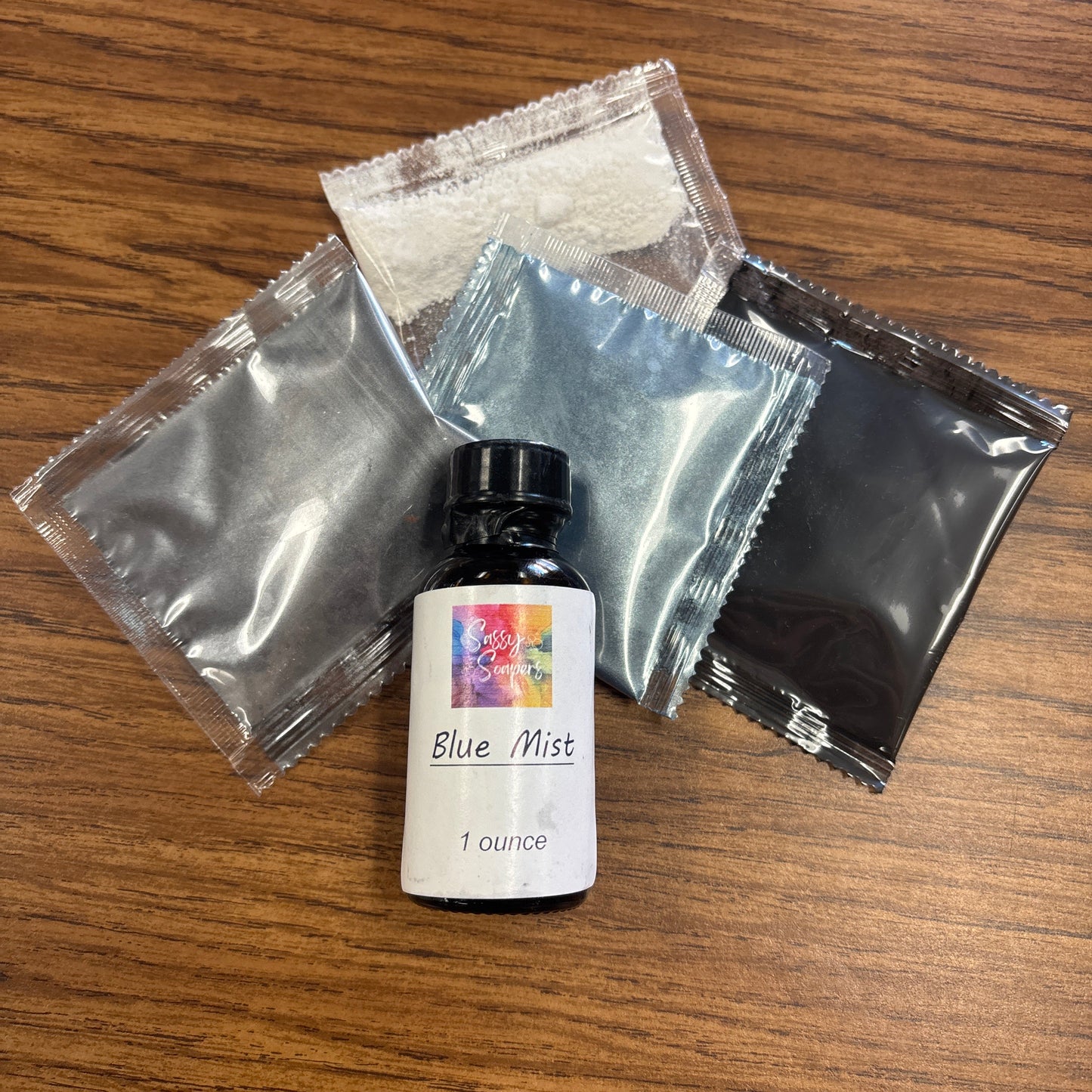 Small bottle with Blue mist fragrence oil label on a wooden surface, surrounded by small packets on a wodden surface 
