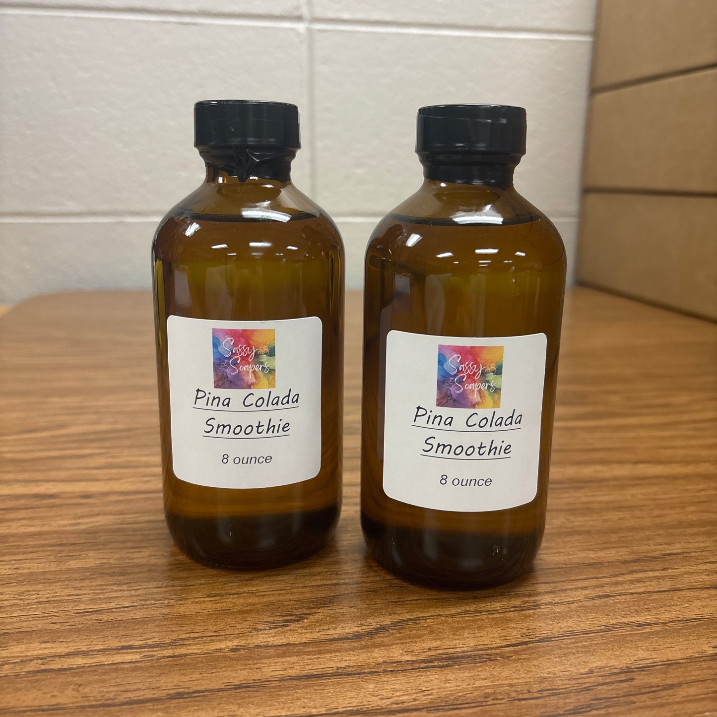Two brown bottles wuth a pina colada smoothie fragrence oils labels on a wooden surface and tiled floor.